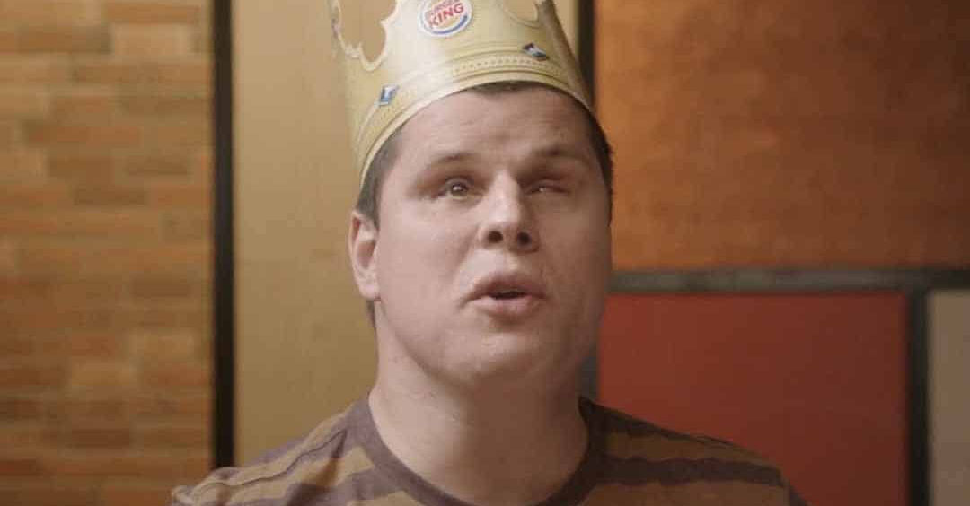 No Burger King, um rapaz cego usa coroa de cartolina, uma camisa listrada marrom e amarelo e segura um sanduíche.