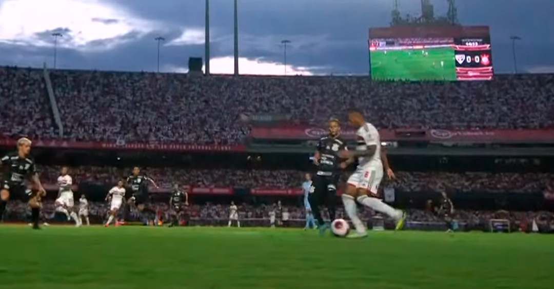 A vista lateral de um campo de futebol com jogadores do São Paulo e do Corinthians disputando a bola. Atrás deles, um telão mostra imagens aéreas do mesmo jogo com o placar marcando zero a zero.