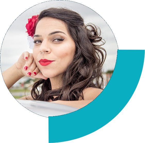 Foto ao ar livre de Daise Alves, ela é uma mulher branca com os cabelos castanhos ondulados em um penteado com duas flores, vermelha e branca, no lado direito. Ela tem sobrancelhas longas e finas, olhos estreitos bem delineados, piercing de argola no septo, batom vermelho. Ela está com a cabeça apoiada em uma das mãos.